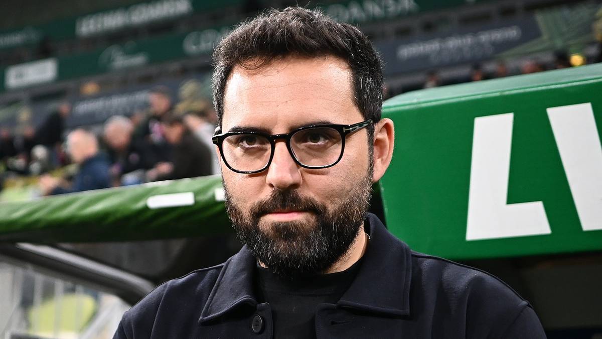 Mężczyzna w okularach przeciwsłonecznych, z brodą, w czarnym swetrze, stoi na tle zielonych siedzeń stadionu.