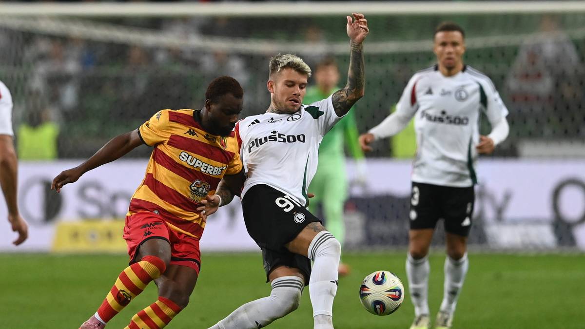 Piłkarz w białej koszulce i czarnych spodenkach walczy o piłkę z piłkarzem w żółto-czerwonej koszulce i czerwonych spodenkach. W tle widać innych piłkarzy.