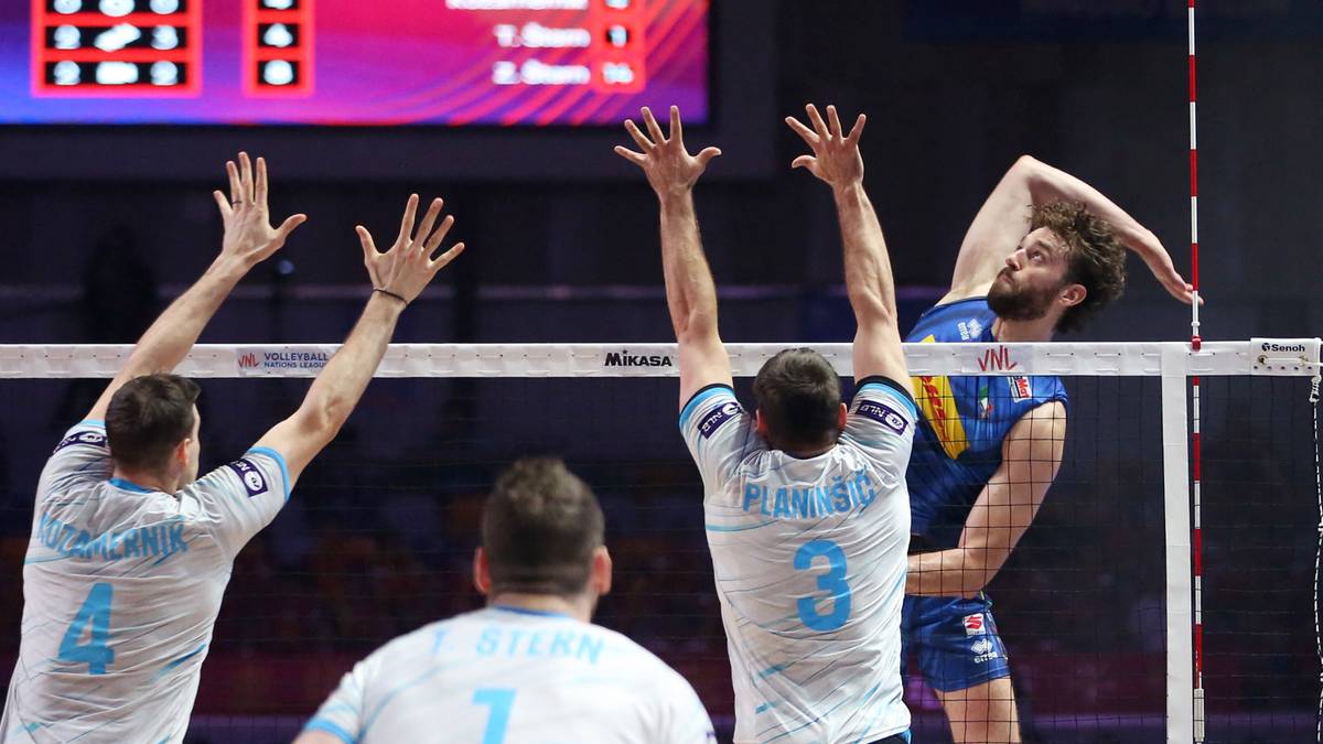 Siatkarze blokują atak przeciwnika podczas meczu. Gracz w niebieskiej koszulce wyskakuje do ataku, podczas gdy dwaj gracze w białych koszulkach próbują go zatrzymać przy siatce.