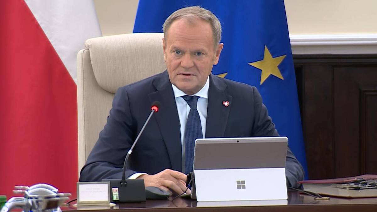 Premier Donald Tusk siedzi przy stole, przed nim laptop, za nim flagi Polski i Unii Europejskiej.