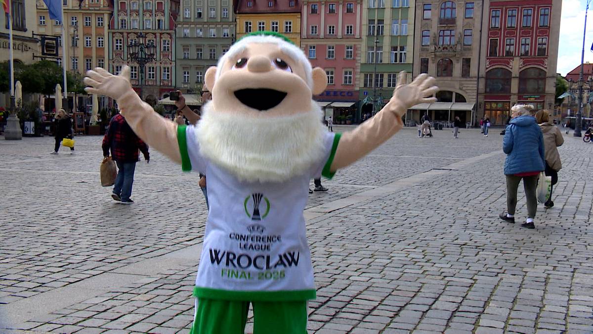 Maskotka w stroju zielono-białym z brodą, trzymająca ręce szeroko otwarte, stojąca na brukowanym placu przed budynkami.