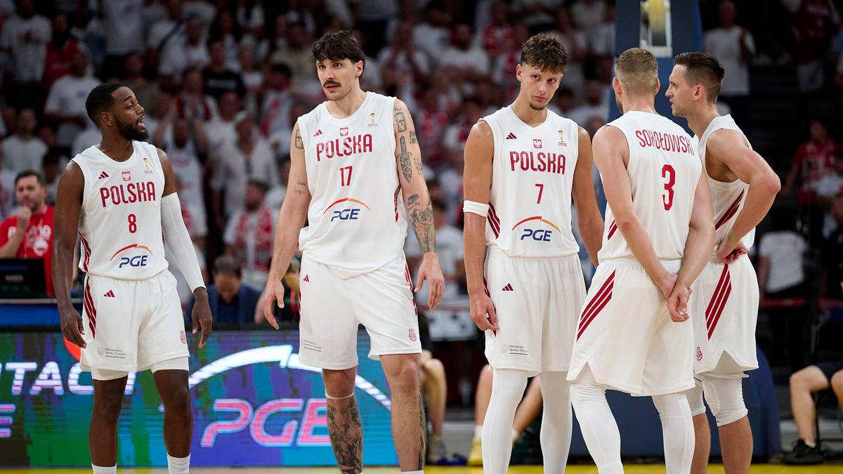Pięciu polskich siatkarzy w białych strojach sportowych z czerwonymi detalami stoi na boisku.