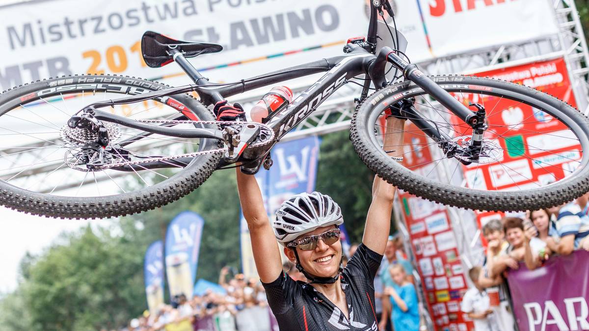 Maja Włoszczowska podnosi ręce w geście triumfu, trzymając kierownicę roweru górskiego.