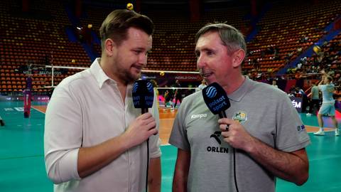 Dw&oacute;ch mężczyzn trzyma mikrofony podczas wywiadu na tle hali sportowej z widownią.