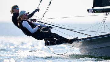 Dwie kobiety w strojach żeglarskich sterują łodzią sportową na wodzie.