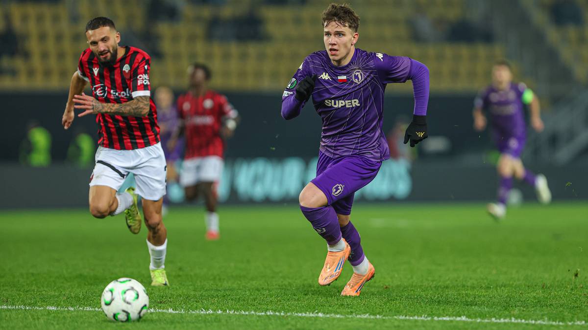 Dw&oacute;ch piłkarzy na boisku, jeden w fioletowym stroju, drugi w pasiastej koszulce, ścigają się z piłką.