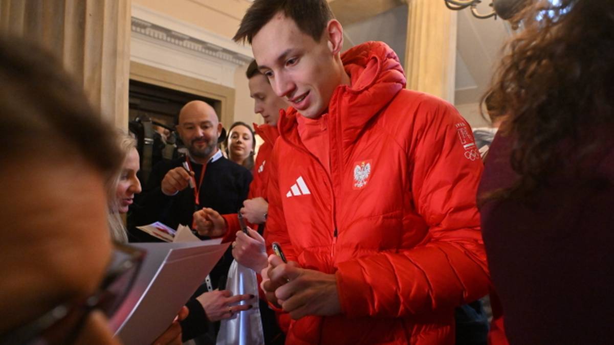 Młody skoczek narciarski Kacper Tomasiak w czerwonej kurtce z godłem Polski rozdaje autografy fanom, otoczony przez tłum.