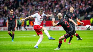 Piłkarze w białych i czarnych strojach walczą o piłkę na boisku piłkarskim, z kibicami na trybunach w tle.