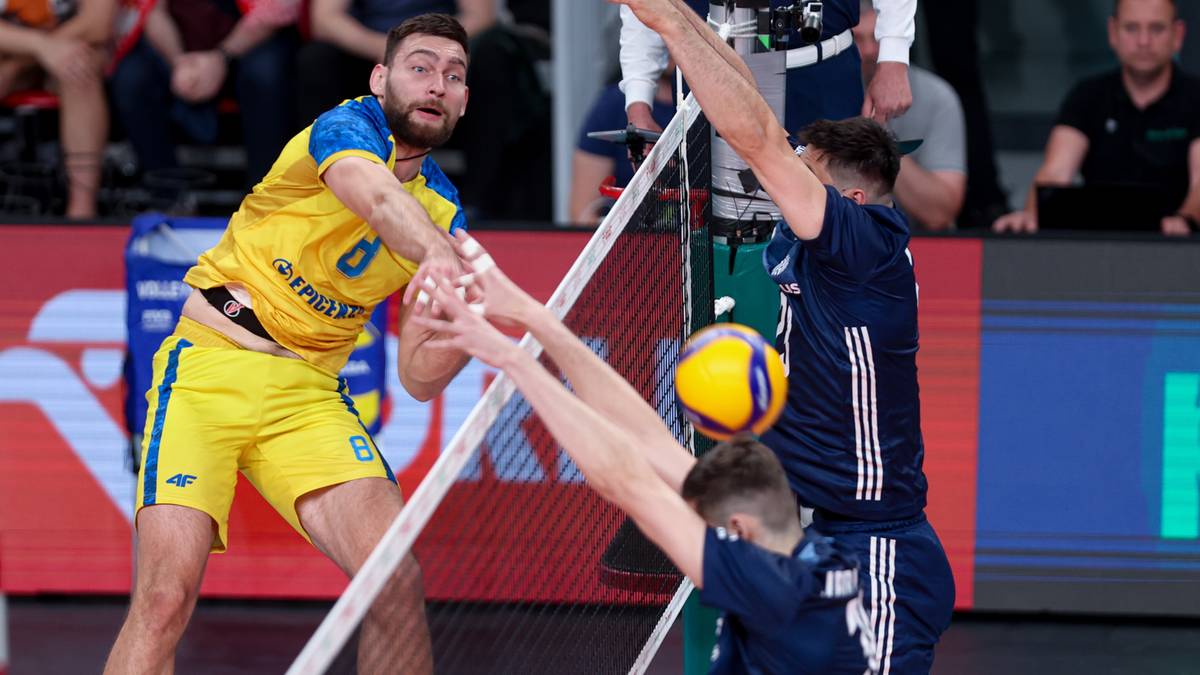 Siatkarz w ż&oacute;łtym stroju pr&oacute;buje zagrać piłkę, blokowany przez przeciwnika przy siatce.