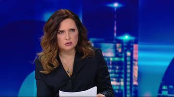 Kobieta w czarnej marynarce i naszyjnikiem, siedzi przy stole z kartką papieru w ręku. Tło stanowi niebieskie studio telewizyjne z sylwetkami budynk&oacute;w.