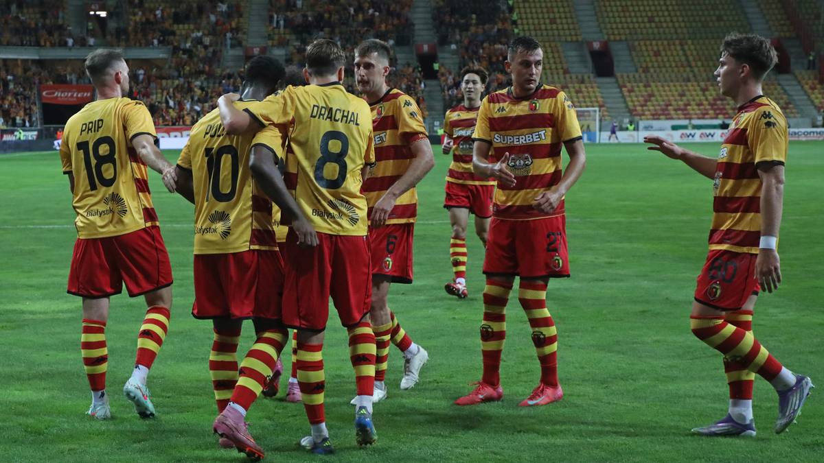 Grupa piłkarzy w żółto-czerwonych strojach sportowych, z numerami na plecach, stoi na zielonej murawie stadionu.