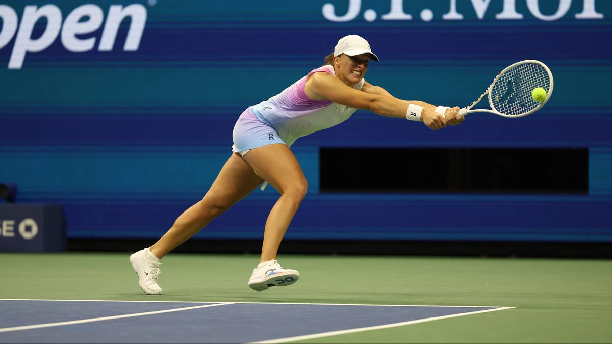 Iga Świątek w akcji na korcie tenisowym, w białej czapce, białej koszulce z kolorowymi pasami i niebieskich spodenkach, odbija piłkę rakietą.