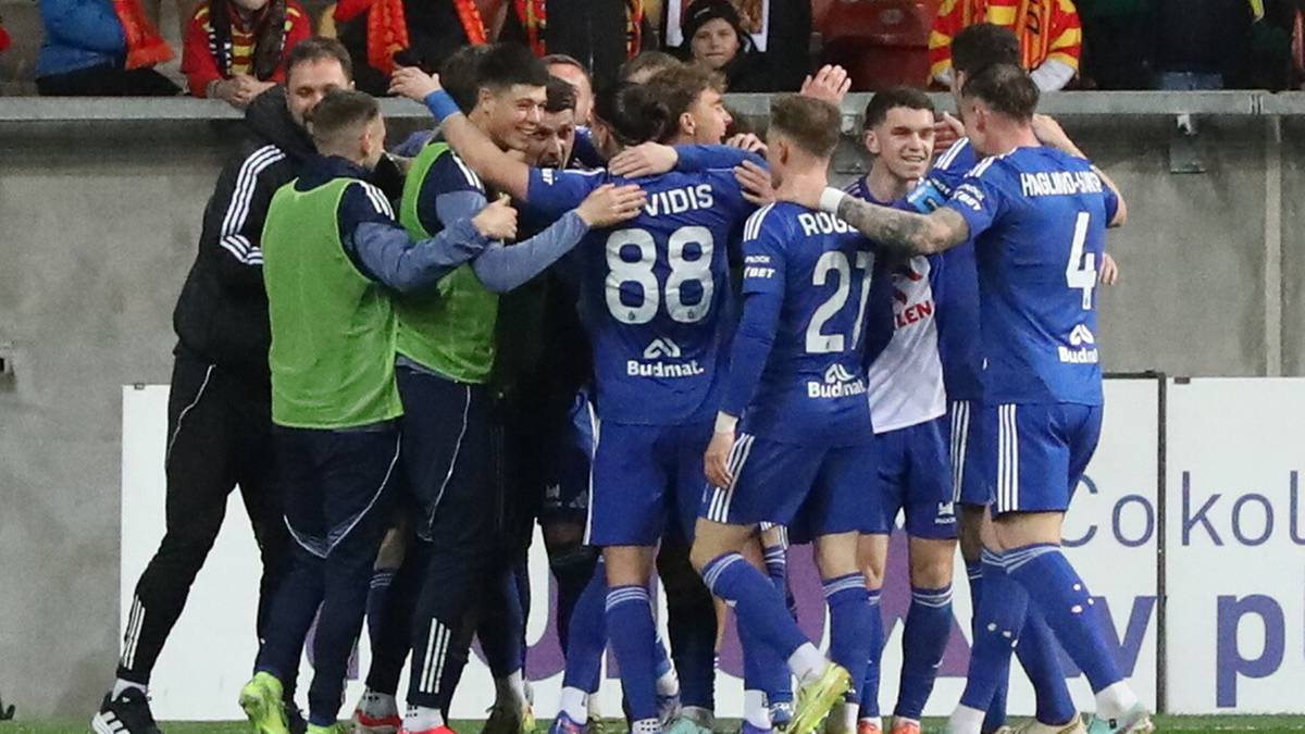 Drużyna piłkarska w niebieskich strojach celebruje razem na boisku, z trybunami kibic&oacute;w w tle.