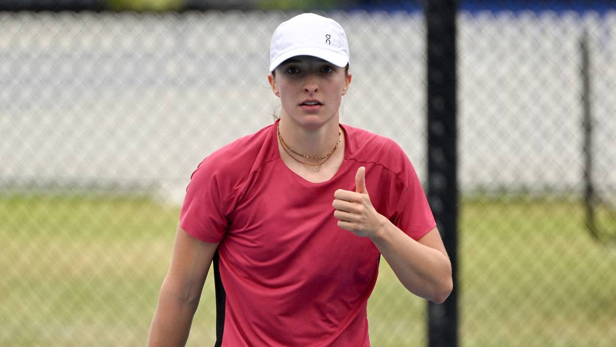 Tenisistka w różowym stroju sportowym i białej czapce idzie po korcie tenisowym, trzymając rakietę i pokazując kciuk w górę.