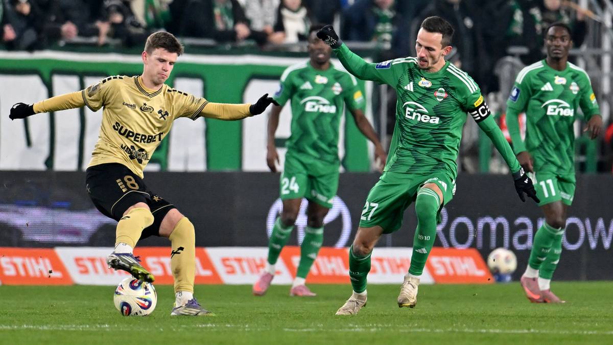 Piłkarz w żółto-czarnym stroju z numerem 18 strzela do bramki, podczas gdy dwóch zawodników w zielonych strojach próbuje go zatrzymać. Na drugim planie widoczni są inni gracze i kibice.