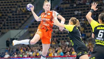 Zawodniczka KGHM Zagłębia Lubin w pomarańczowym stroju, z numerem 91, wykonuje rzut do bramki. Broniąca ją zawodniczka drużyny przeciwnej pr&oacute;buje ją zablokować.