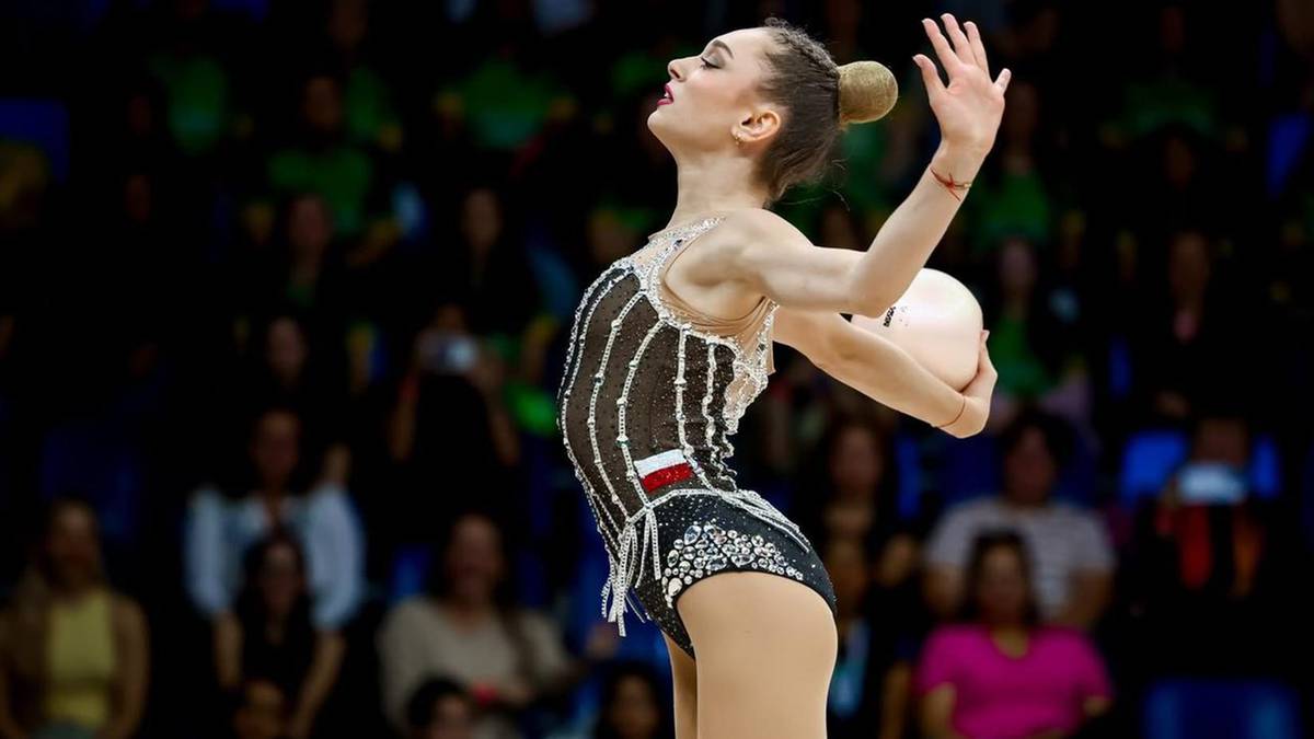 Gimnastyczka artystyczna wykonuje ćwiczenie z piłką na tle publiczności.