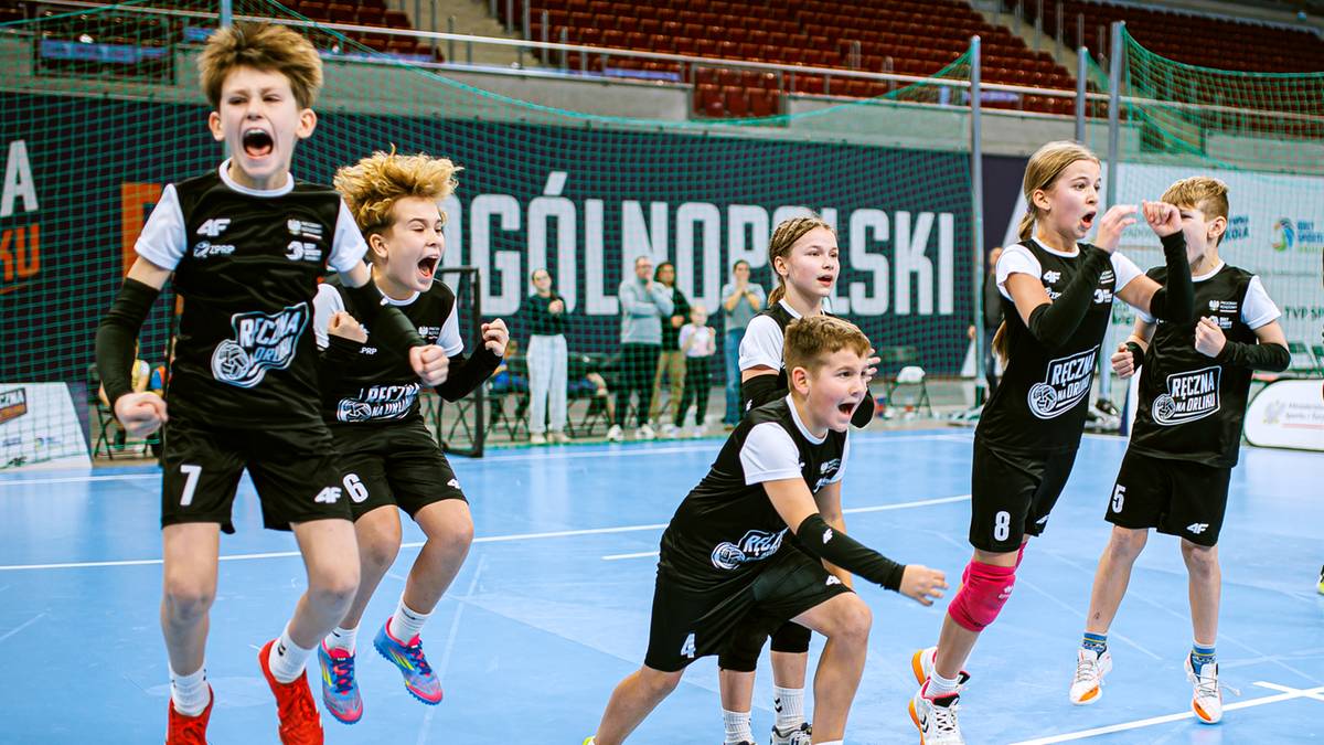 Dzieci w czarnych strojach sportowych cieszą się na boisku do piłki ręcznej, jedne skaczą z radości, inne klęczą.