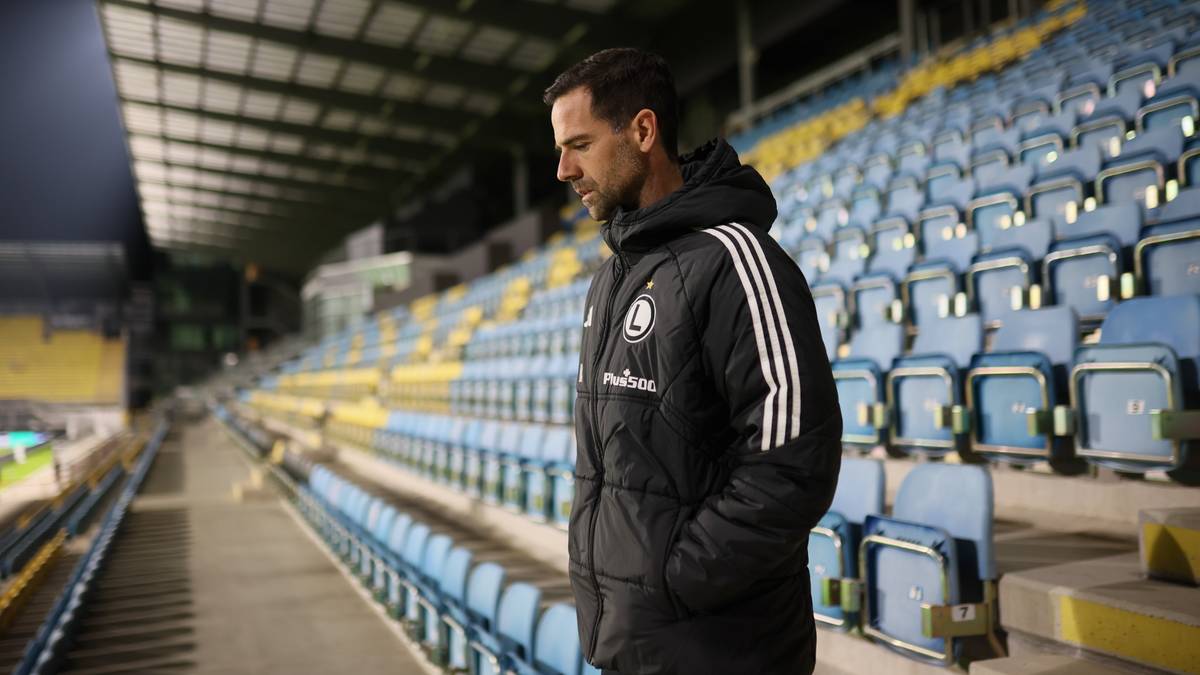 Trener Legii Warszawa w czarnej kurtce stoi na tle pustych trybun stadionu.