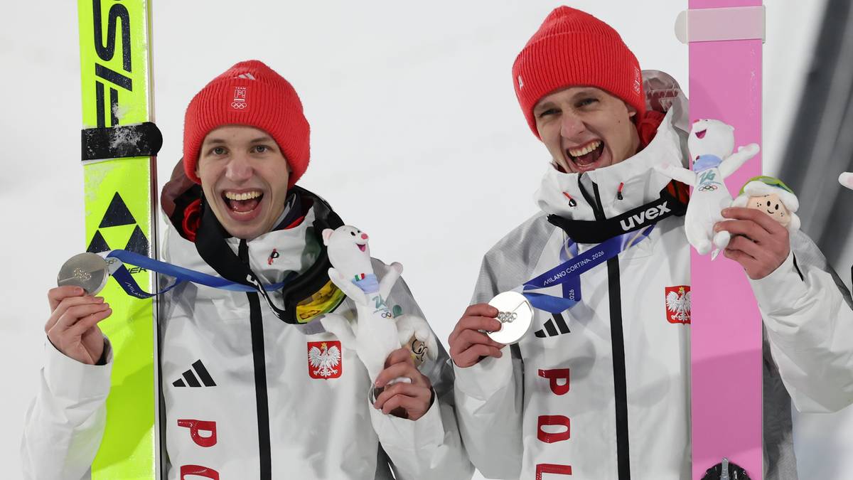 Dwóch polskich skoczków narciarskich w strojach z biało-czerwonymi elementami i czerwonymi czapkami pokazuje medale, stojąc obok nart.