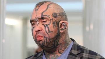 Mężczyzna z licznymi tatuażami na twarzy i szyi udziela wywiadu przed mikrofonem.