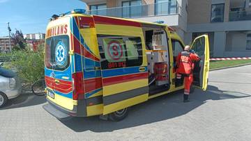 Widok z boku na kolorowy ambulans ratunkowy z otwartymi drzwiami, obok stoi ratownik w czerwonym stroju.