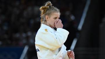 Zawodniczka judo w białym kimonie, z czarnym pasem, zakrywająca usta dłonią, z włosami upiętymi w kok.
