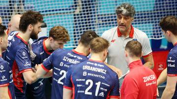 Trener siatk&oacute;wki w białej koszulce m&oacute;wi do grupy zawodnik&oacute;w w niebieskich strojach, kt&oacute;rzy go otaczają i słuchają z uwagą.