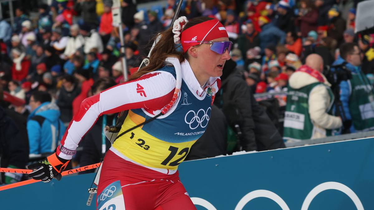 Biathlonistka w czerwonym stroju sportowym z numerem startowym 12, biegnie na trasie zawod&oacute;w biathlonowych. Na plecach ma karabin, a na głowie czapkę i okulary. W tle widoczni są kibice na trybunach oraz niebieska ściana z białymi k&oacute;łkami symbolizującymi igrzyska olimpijskie.