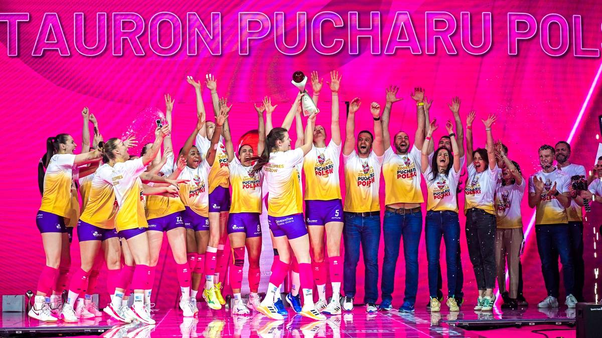Drużyna siatkarek świętuje zwycięstwo, wznosząc ręce i trzymając puchar, na tle r&oacute;żowego banera z napisem Tauron Pucharu Polski.
