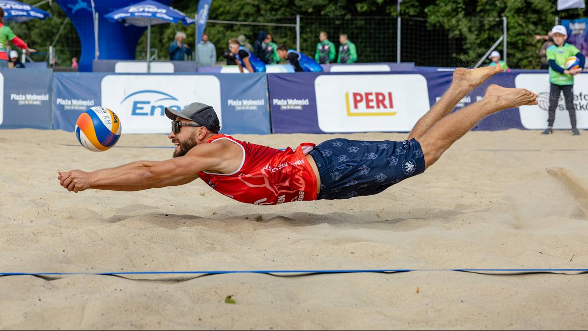 Siatkarz plażowy w białej koszulce i granatowych spodenkach nurkuje, by przyjąć piłkę. Piłka leci w jego kierunku z lewej strony. W tle widać trybuny i inne elementy stadionu.