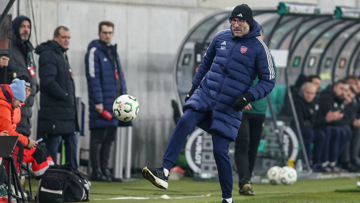 Trener w zimowej kurtce i czapce kopie piłkę na boisku.