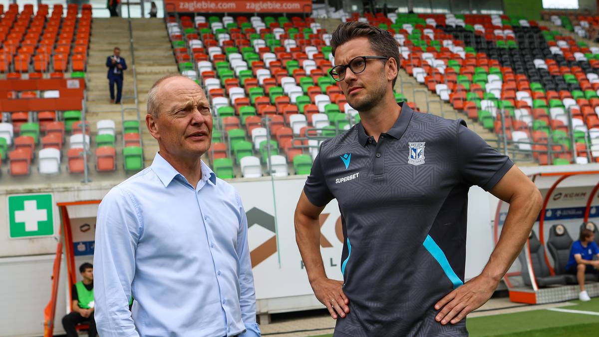 Dw&oacute;ch mężczyzn w strojach sportowych stoi na boisku piłkarskim przed trybunami.