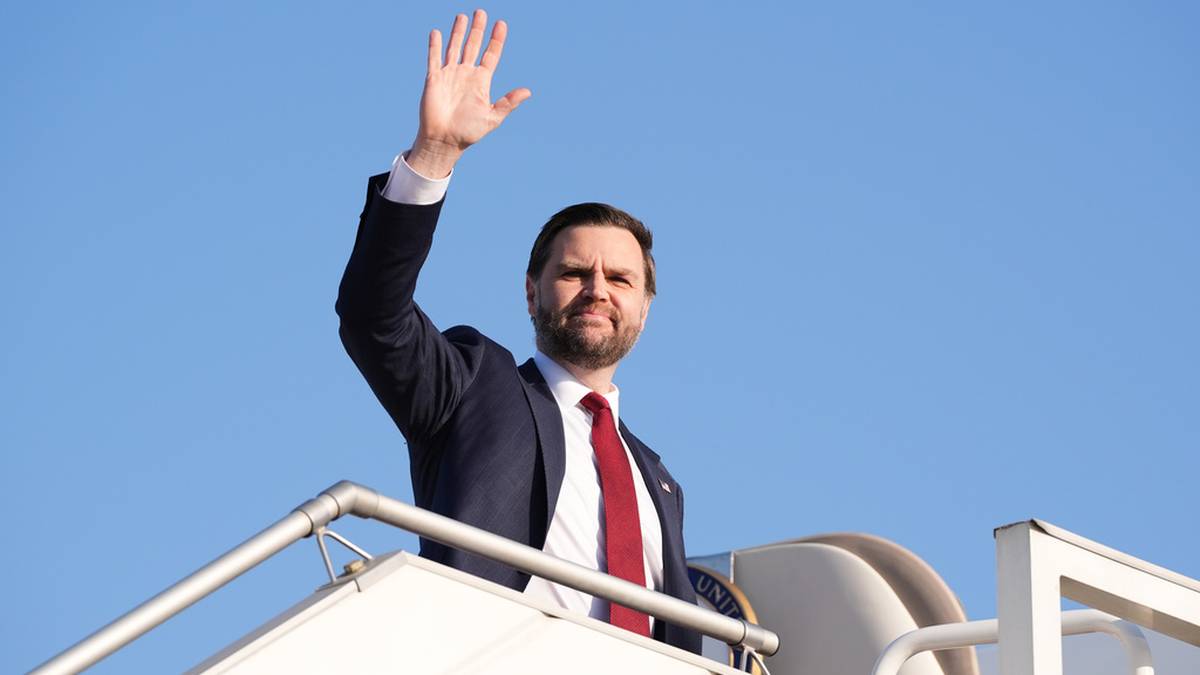 Mężczyzna w garniturze macha prawą ręką na tle błękitnego nieba.