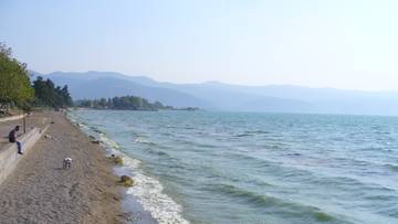 Widok na spokojne, błękitne jezioro z piaszczystą plażą wzdłuż brzegu. W oddali widoczne są łagodne, zalesione wzgórza.