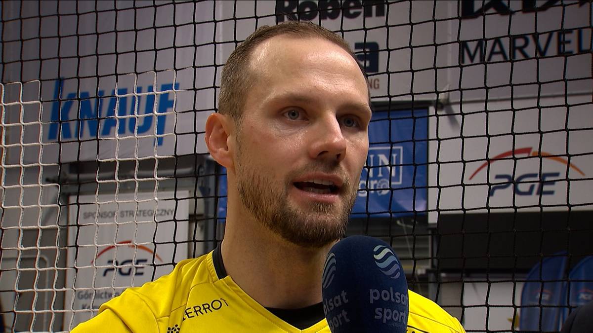 Portret mężczyzny w żółtej koszulce, udzielającego wywiadu. Mężczyzna ma krótką brodę, łysiejącą głowę i patrzy na rozmówcę. W tle widoczna jest siatka oraz banery sponsorów.