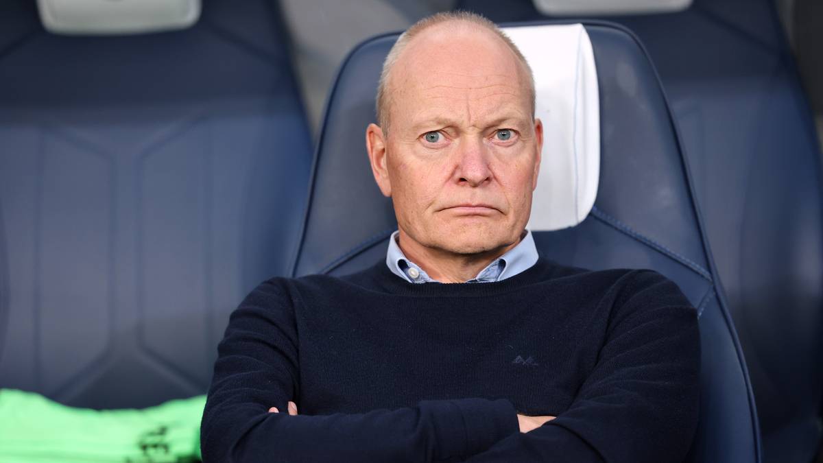 Mężczyzna w średnim wieku, o łysiejącej głowie i surowym wyrazie twarzy, siedzi na krześle z rękami skrzyżowanymi na piersi. Na sobie ma granatowy sweter i niebieską koszulę. Tło stanowi ciemnoniebieska tapicerka siedzeń.