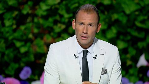 Łukasz Kubot w białym garniturze i czarnym krawacie, w tle zielone liście i fioletowe kwiaty.