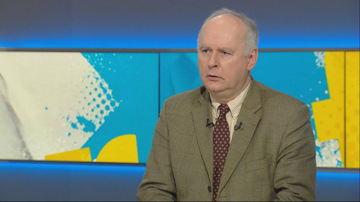 Mężczyzna w beżowym garniturze i krawacie siedzi przed ekranem z kolorowym, abstrakcyjnym wzorem.