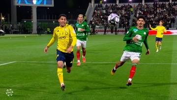 Dwóch piłkarzy w żółtej i zielonej koszulce biegnie po boisku piłkarskim, w tle inni zawodnicy i trybuny.