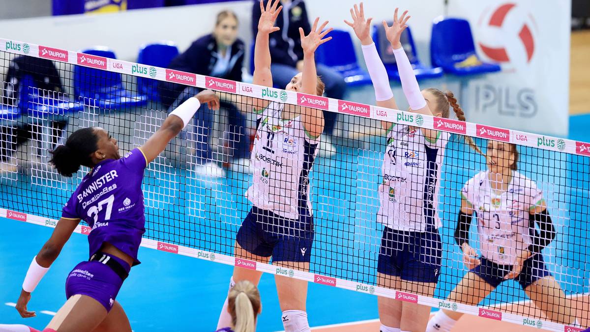 Siatkarka w fioletowym stroju atakuje piłkę nad siatką, podczas gdy inne zawodniczki z drużyny przeciwnej pr&oacute;bują ją zablokować.