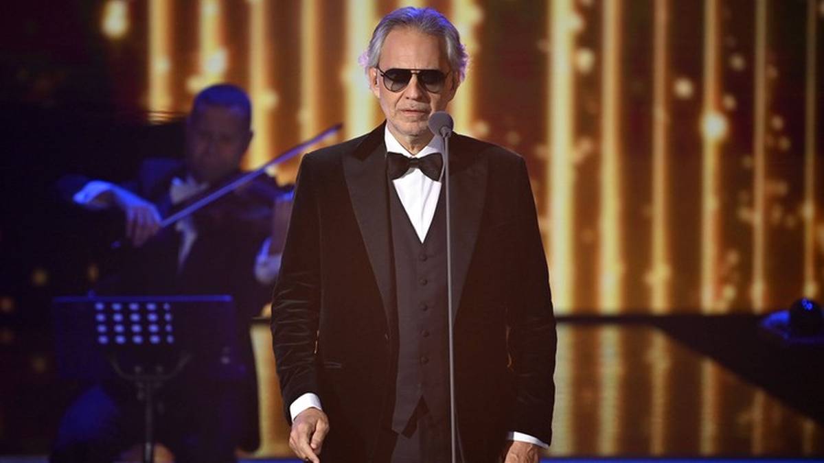 Andrea Bocelli w czarnym smokingu i okularach przeciwsłonecznych stoi na scenie, śpiewając do mikrofonu. W tle gra skrzypek.