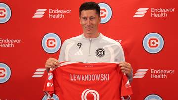 Robert Lewandowski trzyma czerwono-białą koszulkę piłkarską z napisem "LEWANDOWSKI" na plecach, stojąc przed ścianką z logo "Endeavor Health" i symbolem "C" w okręgu.