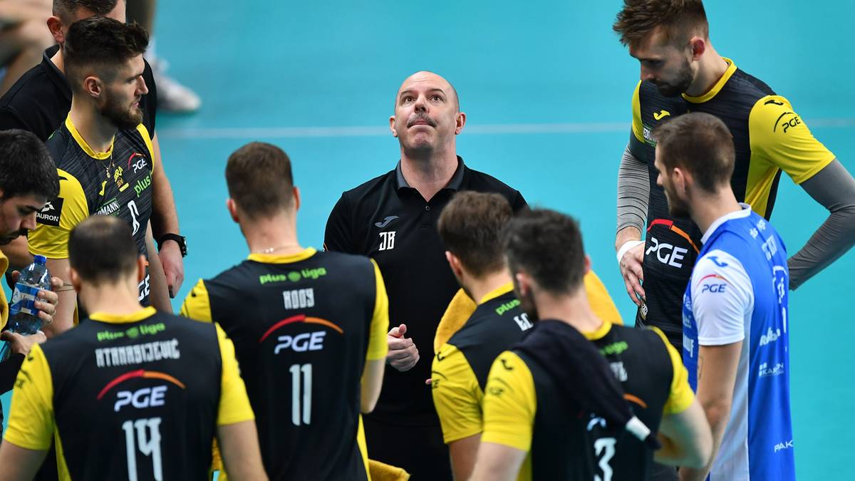 Trener siatk&oacute;wki przemawia do grupy zawodnik&oacute;w w czarnych strojach sportowych.