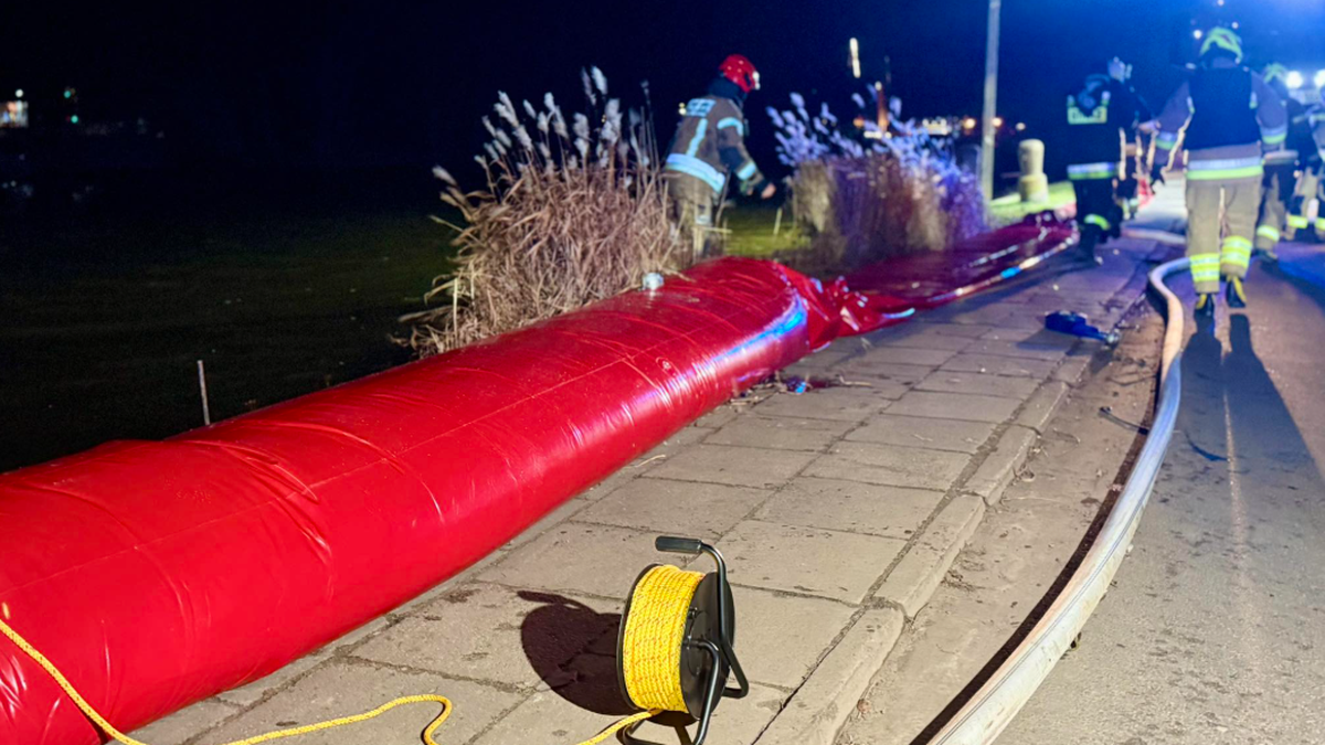 Strażacy rozwijają czerwony wąż lub barierę przeciwpowodziową wzdłuż nabrzeża nocą, w tle widać światła pojazd&oacute;w.