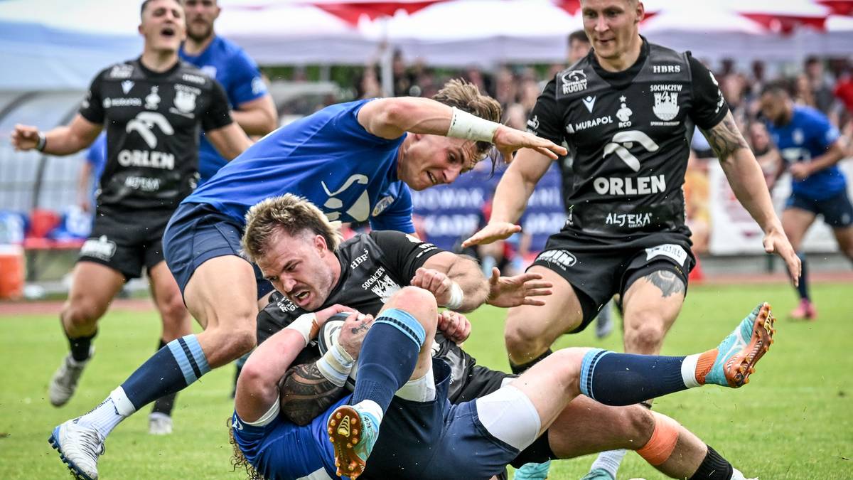 Rugbiści walczą o piłkę na boisku, jeden z zawodnik&oacute;w pr&oacute;buje ją odebrać przeciwnikowi leżącemu na ziemi.