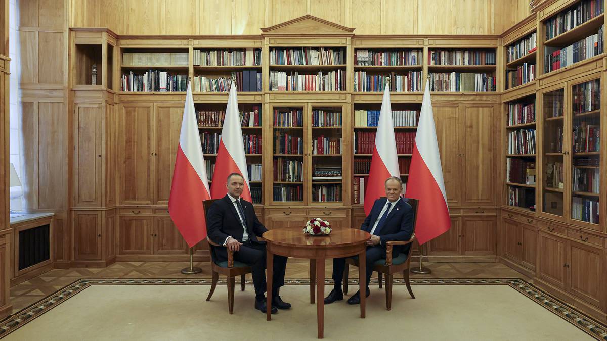 Dw&oacute;ch mężczyzn w garniturach siedzi przy okrągłym stole, z polskimi flagami po bokach, w pomieszczeniu z regałami pełnymi książek.