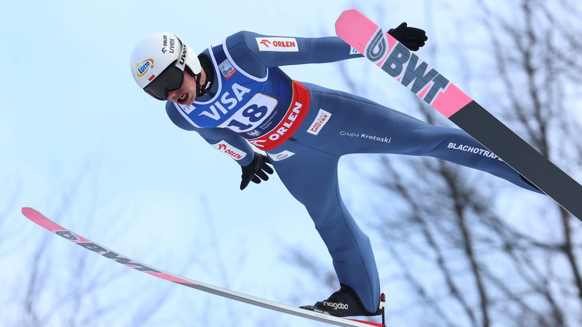 Skoczek narciarski w niebieskim kombinezonie i białym kasku wykonujący skok na nartach.