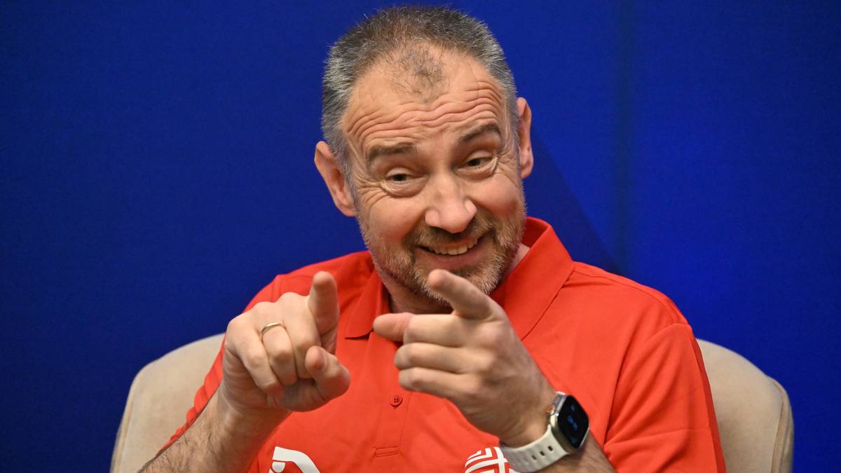 Mężczyzna w czerwonej koszulce z kołnierzykiem, z kr&oacute;tkimi siwymi włosami i kilkudniowym zarostem, uśmiecha się i wskazuje dwoma palcami w stronę widza.