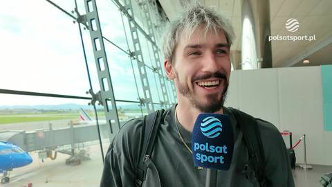 Portret Tomasza Fornala z szerokim uśmiechem, trzymającego mikrofon Polsat Sport. W tle widok na płytę lotniska z samolotem i rękawem.